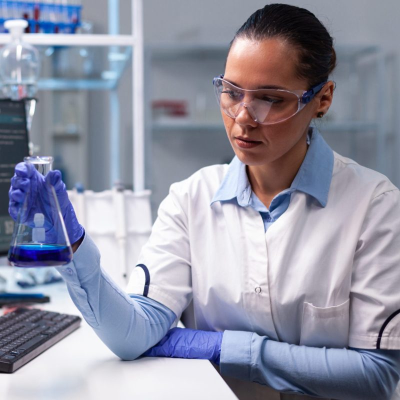 Scientist woman doctor holding glass flask analyzing liquid solution working at biochemistry experiment in microbiology hospital laboratory. Specialist researcher discovering medical diagnostic